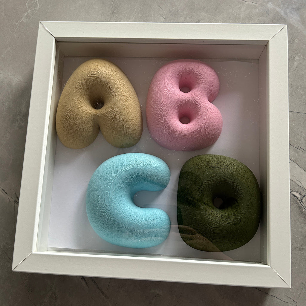 A square white shadow box display containing four soft foam letters and numbers arranged to spell 'ABCD' in a 2x2 grid. The letters and numbers are in pastel shades of beige, pink, light blue, and olive green, set against a white background inside the frame, photographed from above on a gray marble surface.