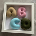 A square white shadow box display containing four soft foam letters and numbers arranged to spell 'ABCD' in a 2x2 grid. The letters and numbers are in pastel shades of beige, pink, light blue, and olive green, set against a white background inside the frame, photographed from above on a gray marble surface.