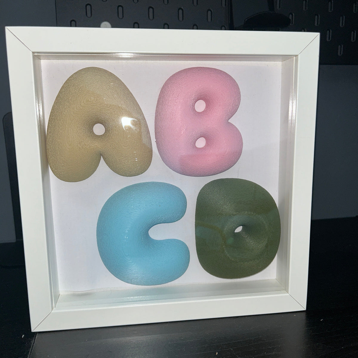A white square shadow box frame displaying four stylized lowercase letters in beige, pink, light blue, and olive green, arranged to spell 'abco' on a white background.