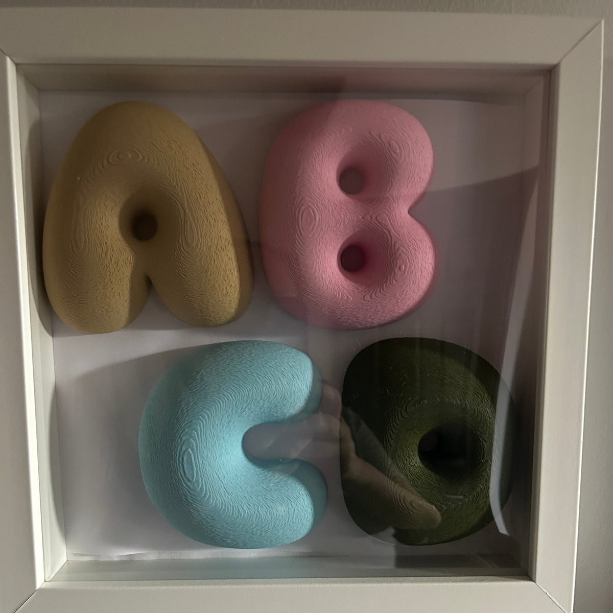 A framed wall art piece showing the letters A, B, C, and D shaped like donuts in muted beige, pink, light blue, and dark olive green, set in a white shadowbox frame against a light grey wall.