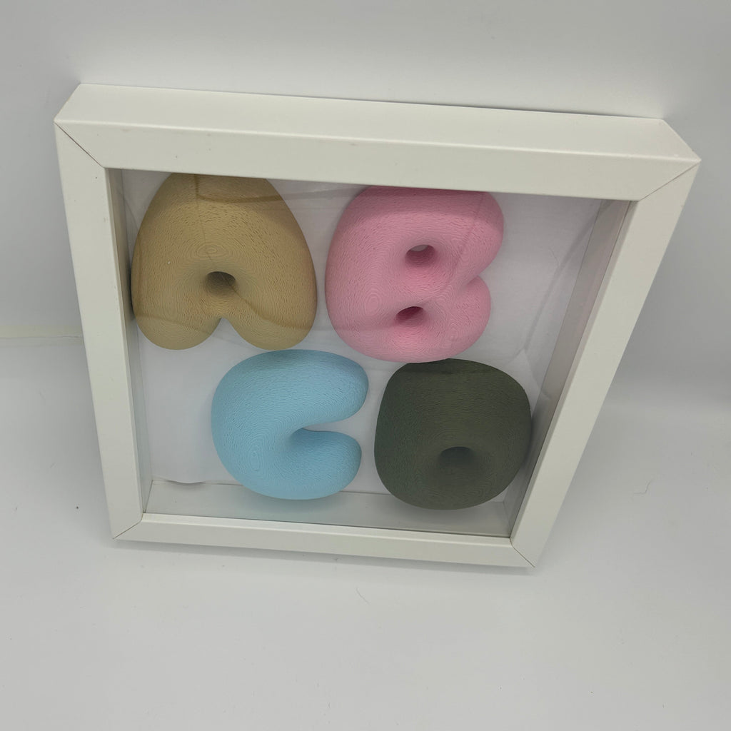 A white square shadow box display containing four soft foam letters spelling 'BUBG' in muted pastel colors: tan, pink, light blue, and olive green, photographed on a white surface.