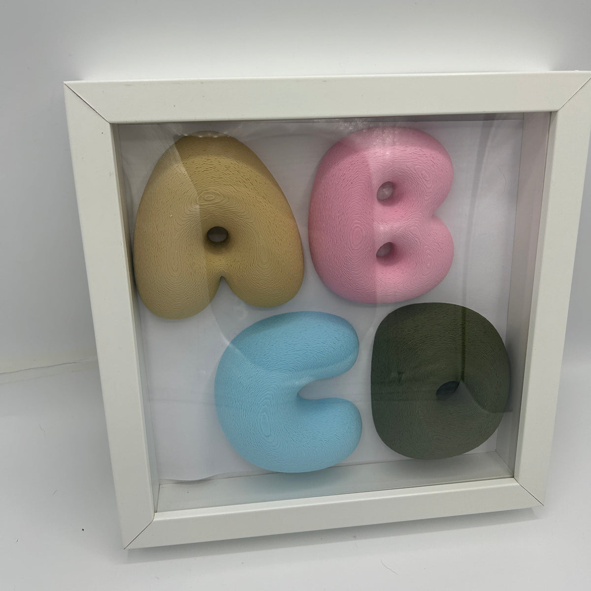 A square white shadow box frame displaying four pastel-colored foam letters arranged in a 2x2 grid: tan, pink, light blue, and dark olive green.