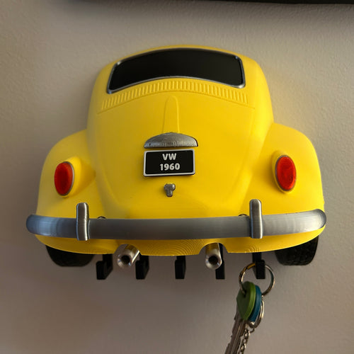 A bright yellow novelty key holder shaped like the rear of a classic VW Beetle, mounted on a light-colored wall with keys hanging from the tailpipe area.