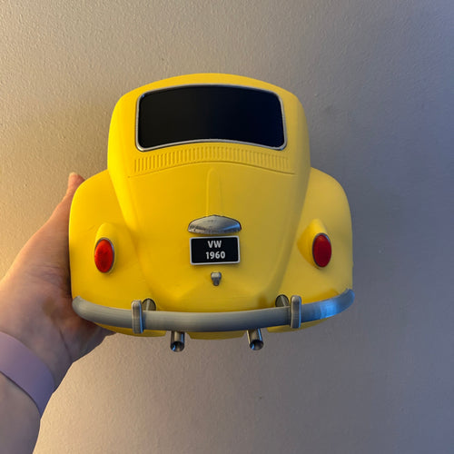 A hand holding a bright yellow Beetle car key holder against a plain beige wall.