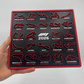 A hand holds a square black F1 2026 wall plaque featuring 20 circuit track outlines in red with white and red text labels, against a plain light gray background.