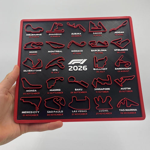 A hand holds a square black F1 2026 wall plaque featuring 20 circuit track outlines in red with white and red text labels, against a plain light gray background.