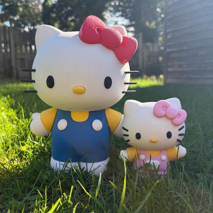 Two Hello Kitty garden statues in outdoor grass, one larger figure wearing blue overalls with a pink bow, and one smaller figure wearing pink polka dots with a pink bow.