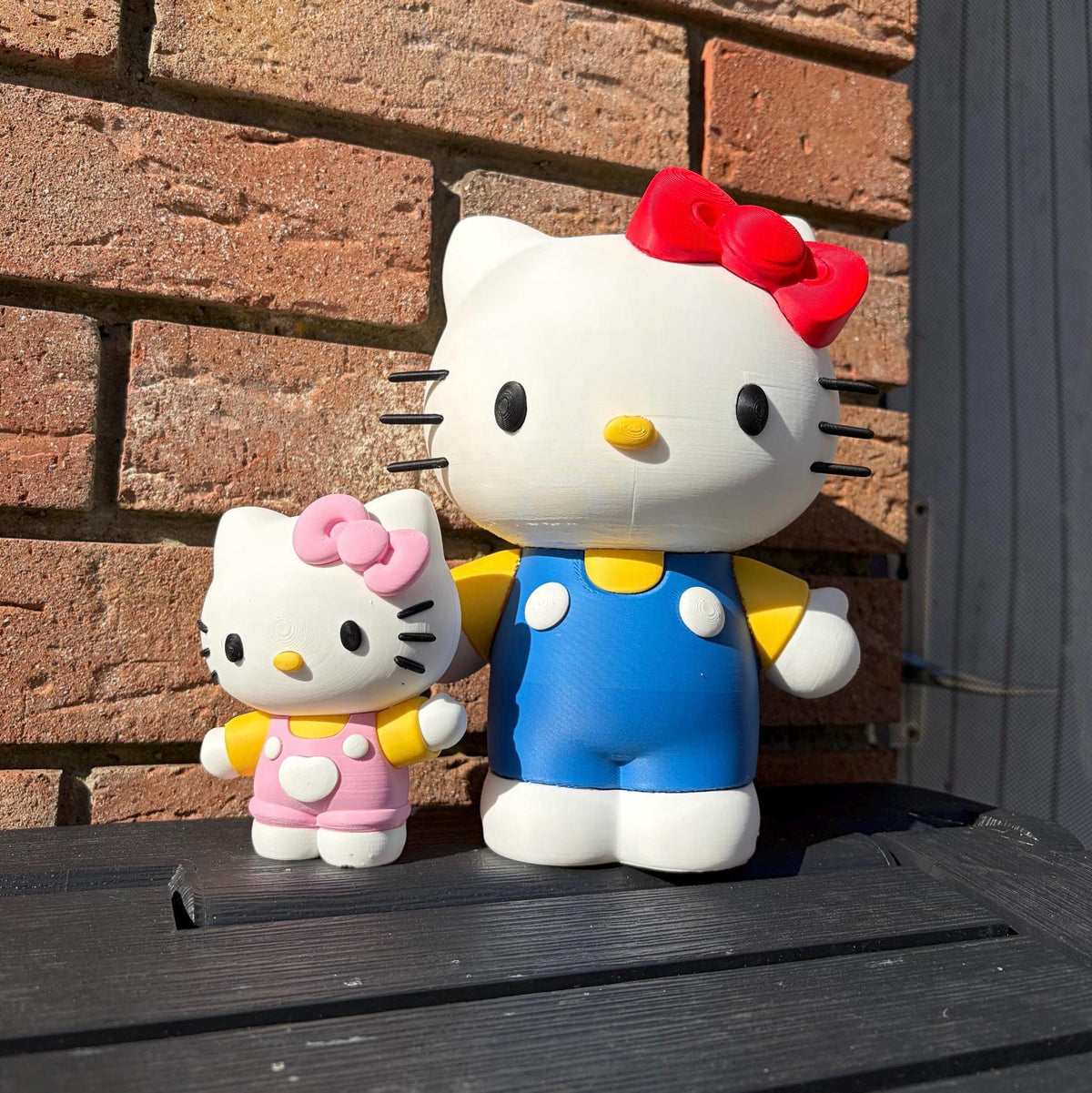Two Hello Kitty figurines, one large and one small, standing on a black wooden surface in front of a red-brown brick wall. The large Hello Kitty figure wears a red bow, blue overalls, and yellow sleeves, while the smaller figure wears a pink bow and pink overalls.