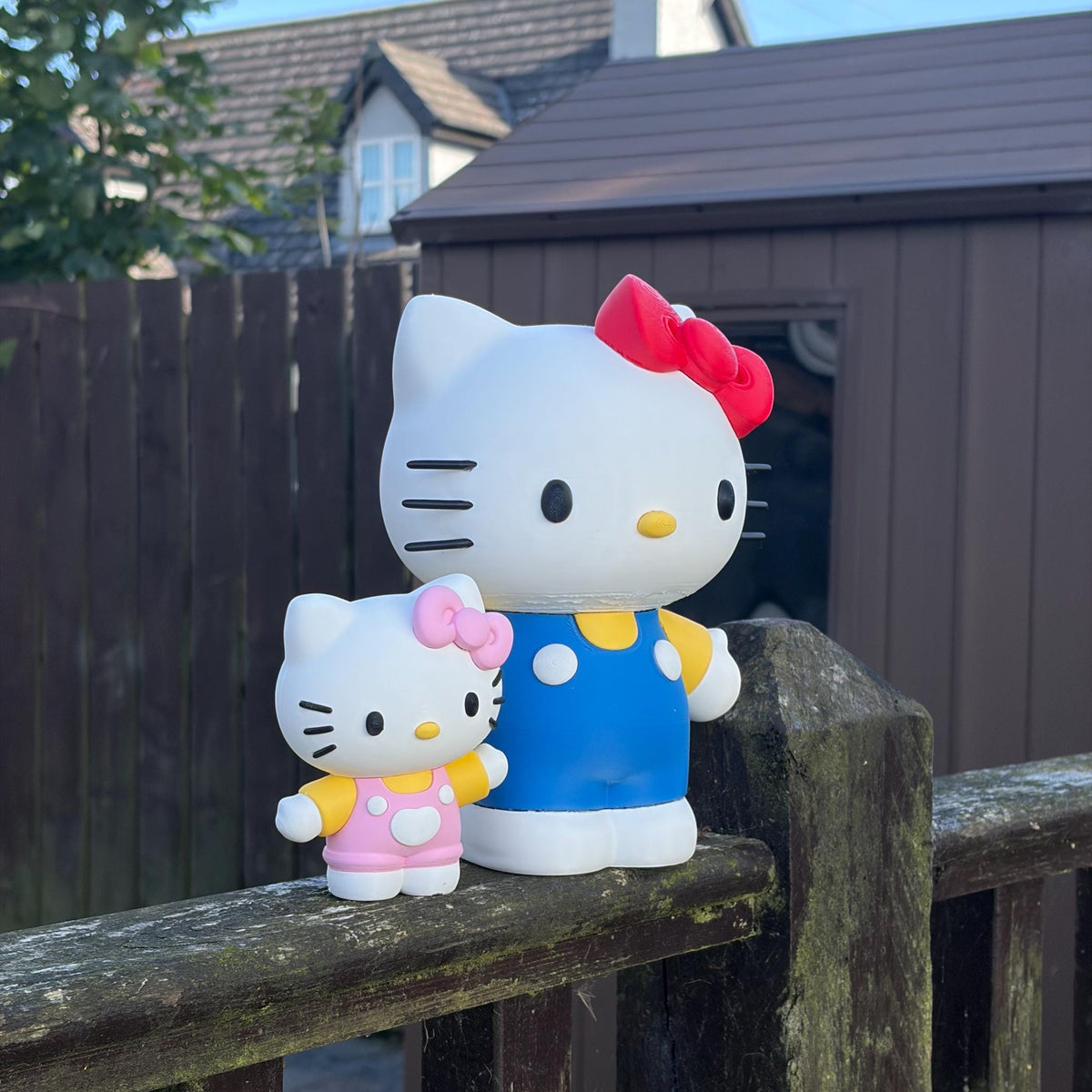 Two Hello Kitty figurines, one large and one small, displayed outdoors on a weathered wooden fence with a brown shed and houses in the background.