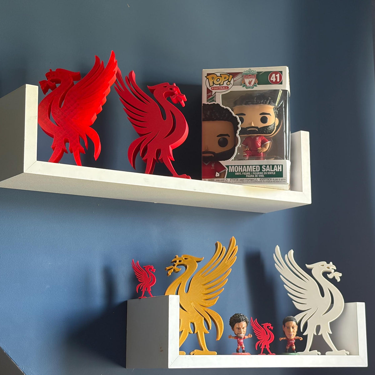 Two white floating shelves mounted on a dark blue wall, styled with red and yellow Liverbird cutouts, a white Liverbird cutout, and a Funko Pop! figure of Mohamed Salah.