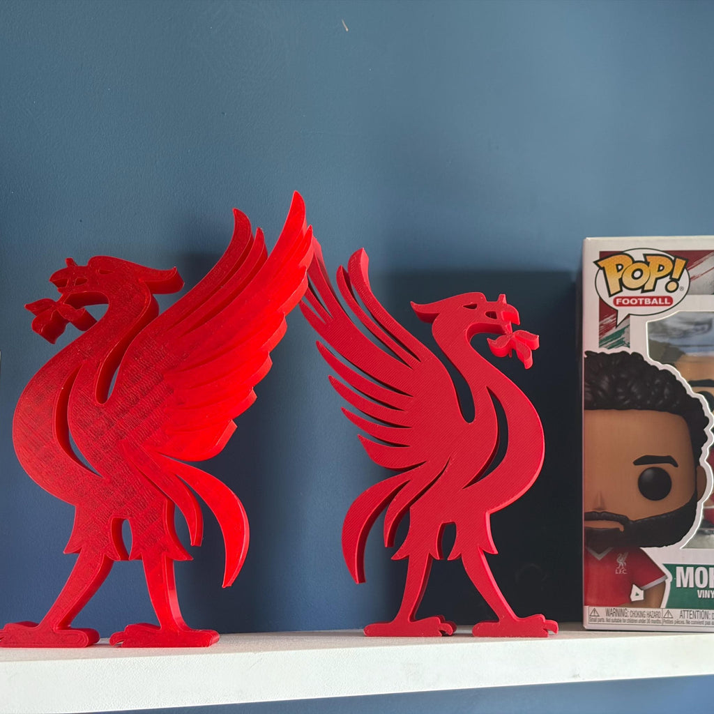 Two red Liverpool bird 3D printed sculptures displayed on a white shelf next to a boxed Funko Pop! Mohamed Salah figure against a dark blue wall.