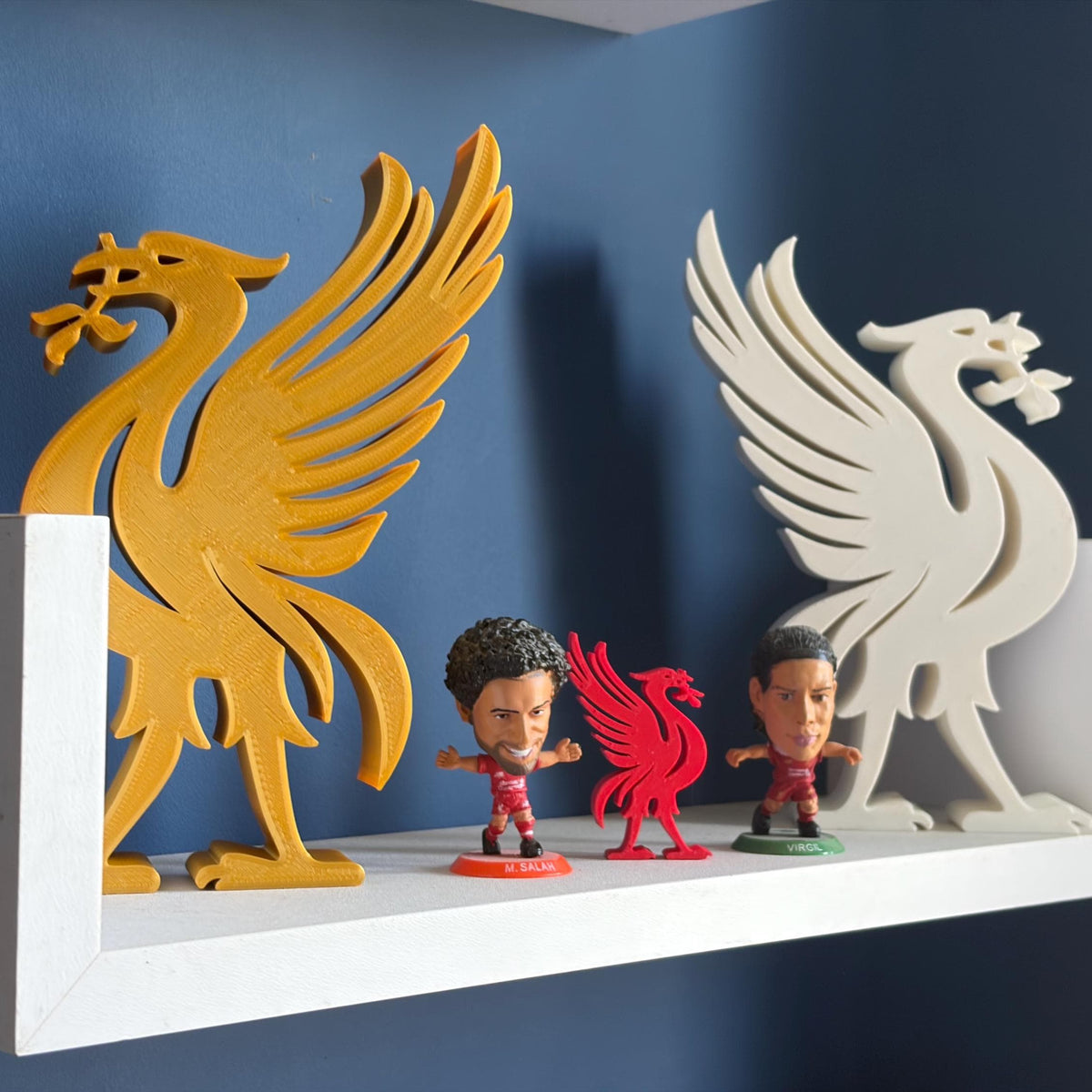 A set of Liverpool FC themed wall decorations displayed on a white floating shelf against a dark blue wall. The set includes a large gold and white 3D prints of the Liverbird, a small red 3D print and two small figurines of Liverpool players.