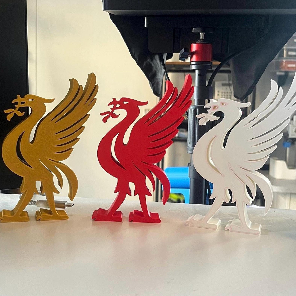 Three 3D printed Liverpool bird figurines in gold, red, and white, displayed on a white surface in a workshop setting.