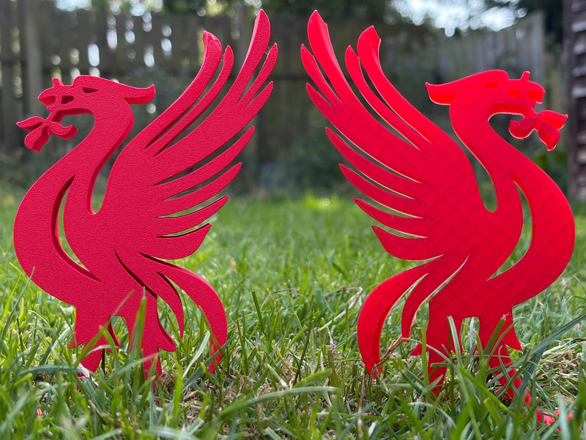 Two red cutout bird garden stakes shaped like Liverpool birds standing in green grass outdoors.