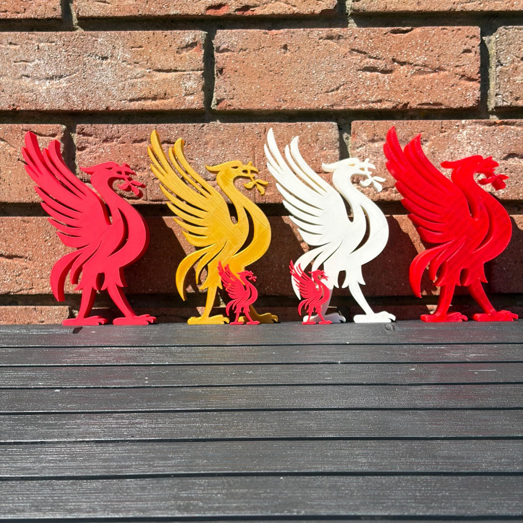 Six Liverpool bird 3D printed decorations in red, yellow, and white, arranged in a row on a dark wooden surface against a brick wall background.