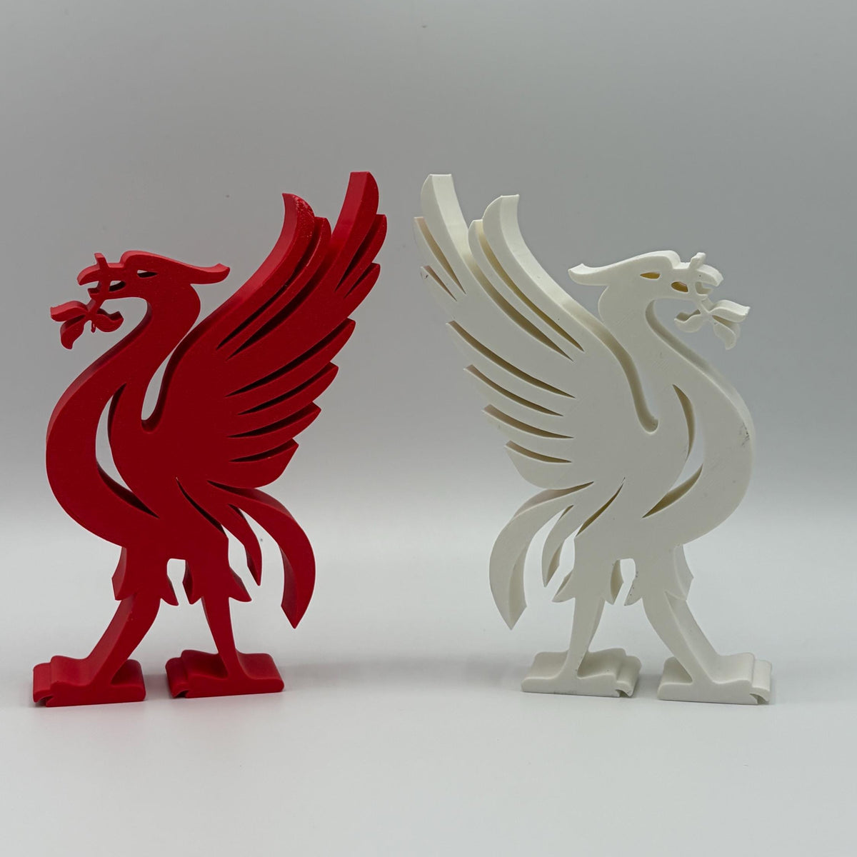 Two Liverbird figurines, one in red and one in white, displayed side-by-side on a light gray background.