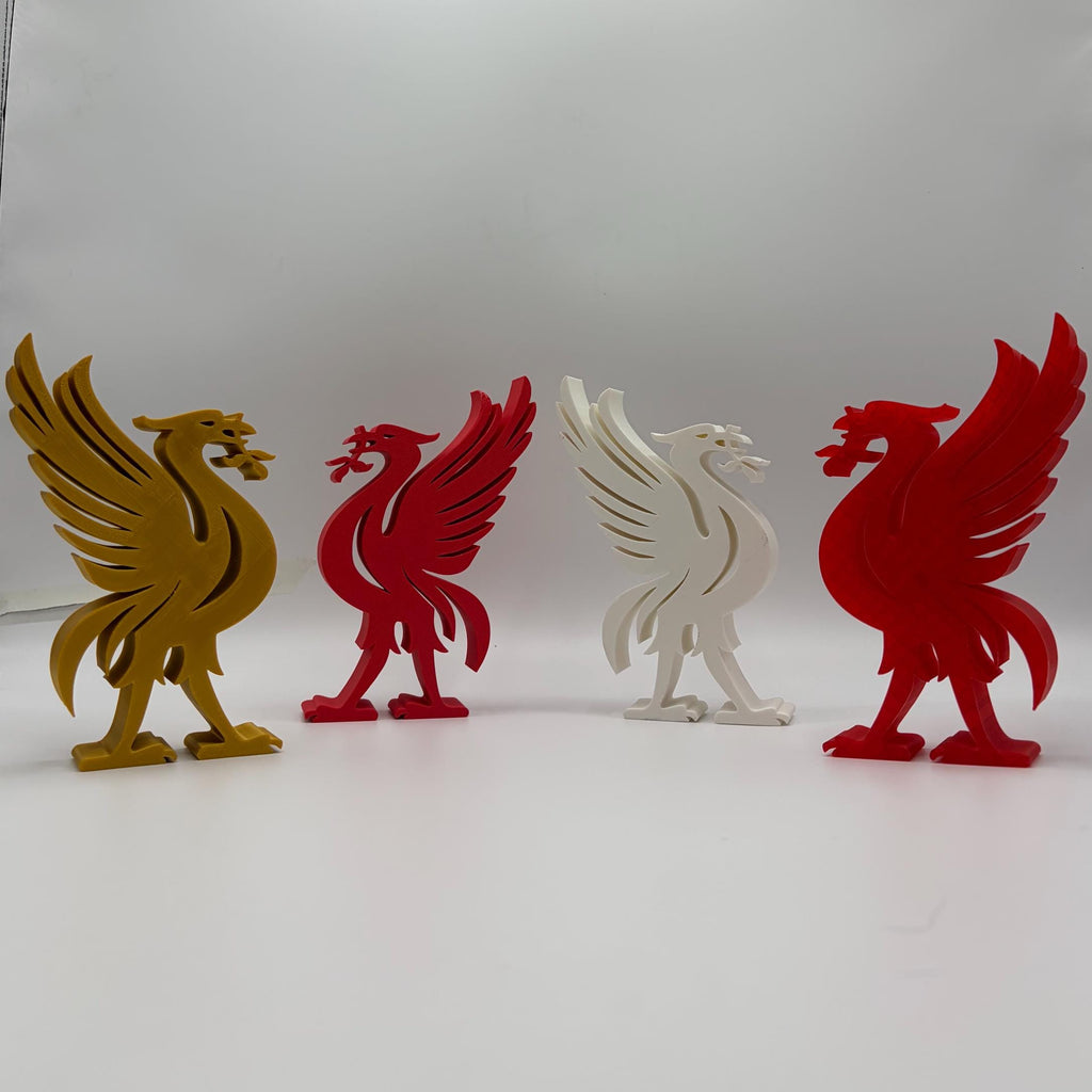 Four 3D-printed cutout figurines of the Liverpool city crest (liver bird) in gold, red, and white, arranged in a row on a white surface inside a lightbox.