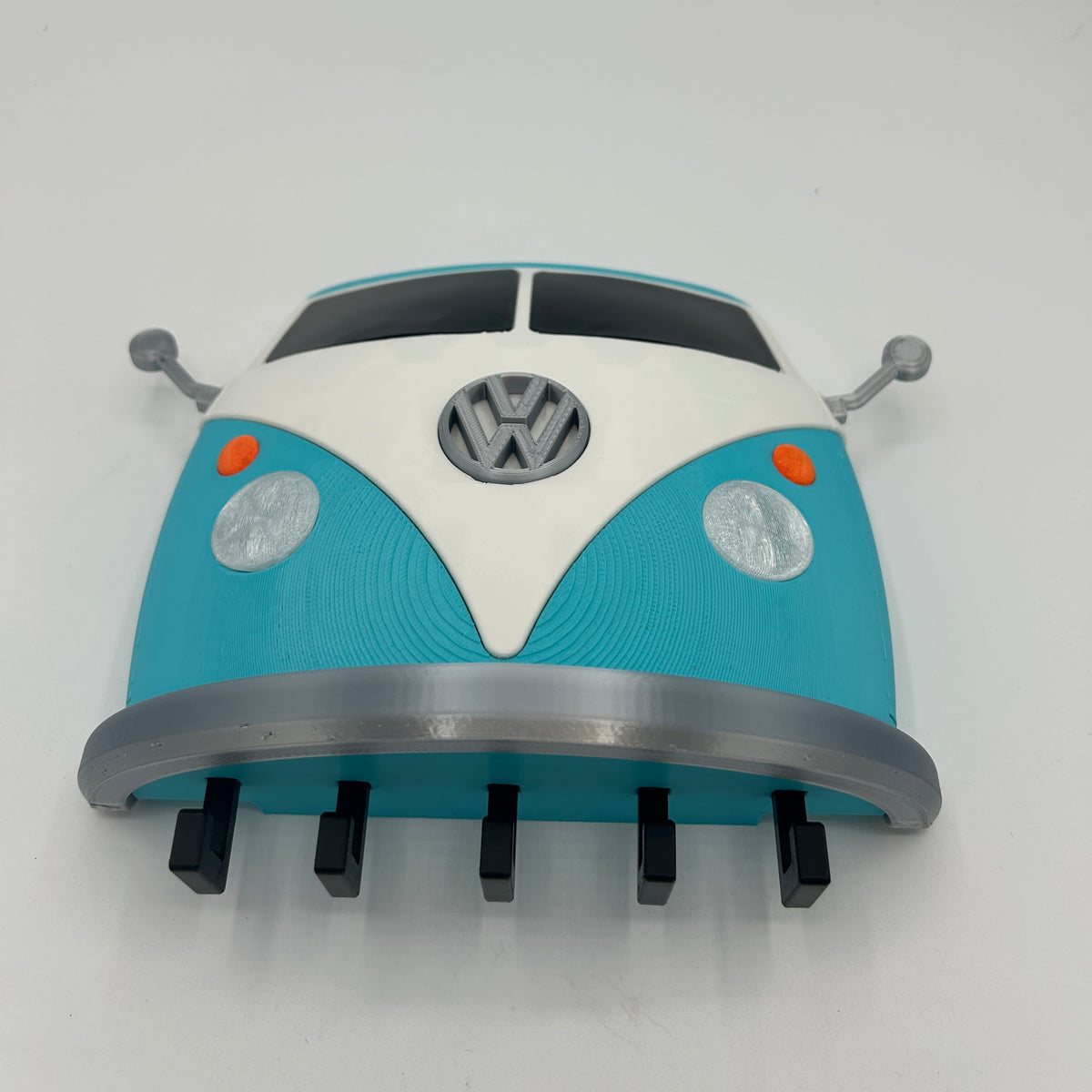 A novelty wall clock shaped like the front of a classic V Dub Type 2 van, featuring a turquoise and white body with a grey logo, black tires, and silver side mirrors, photographed against a plain white background.