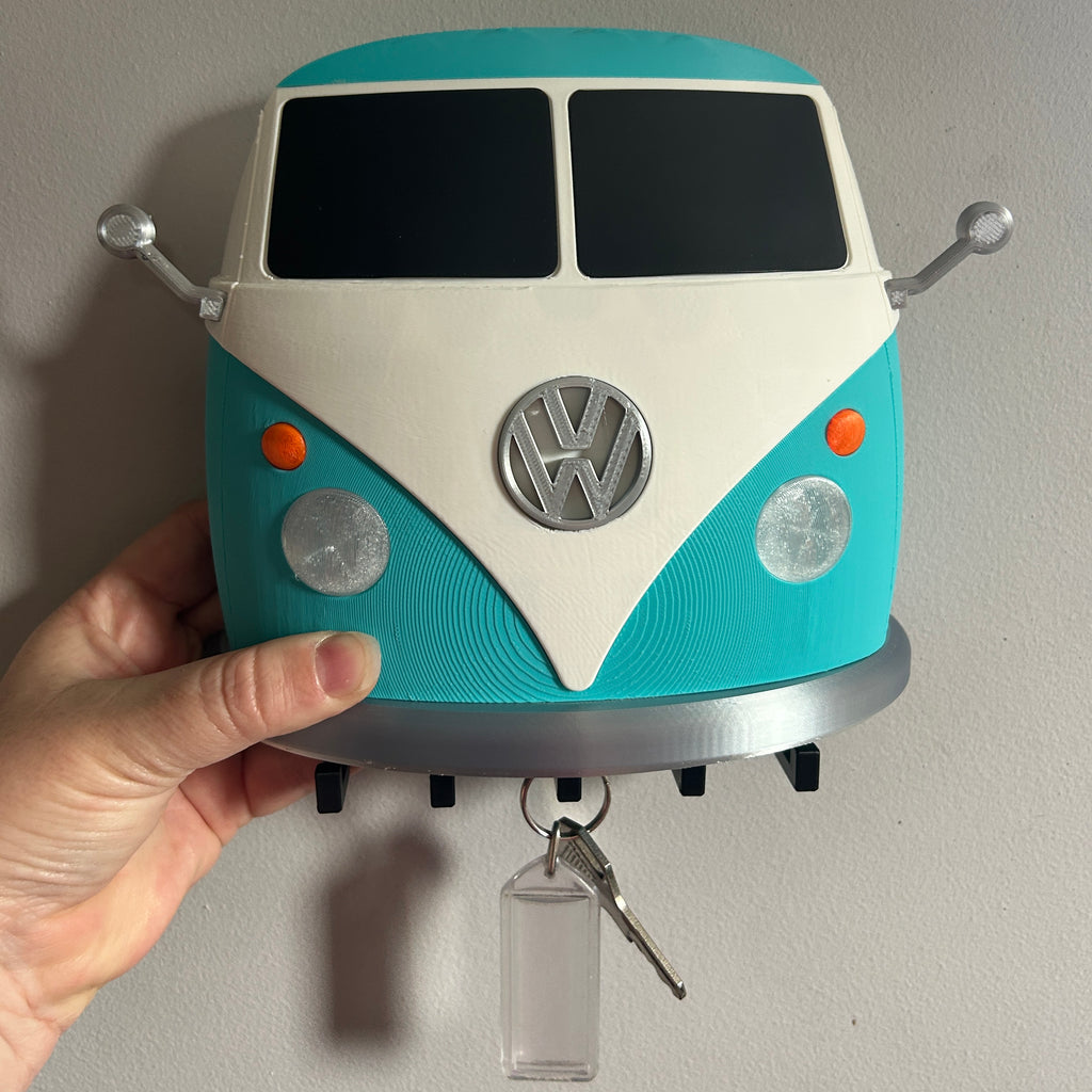 A hand holding a turquoise and cream Volkswagen-style bus-shaped key holder against a plain wall, with keys hanging from the front.