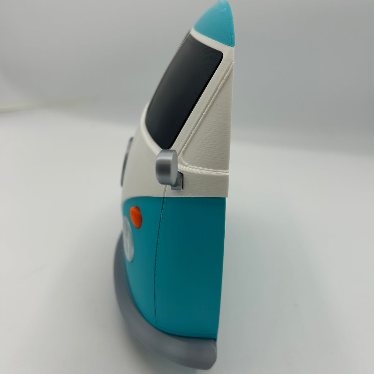 A close-up side view of a retro-style toy vehicle, resembling a miniature toy van, featuring a turquoise blue body, white upper section, dark window, grey base, and small orange accent.