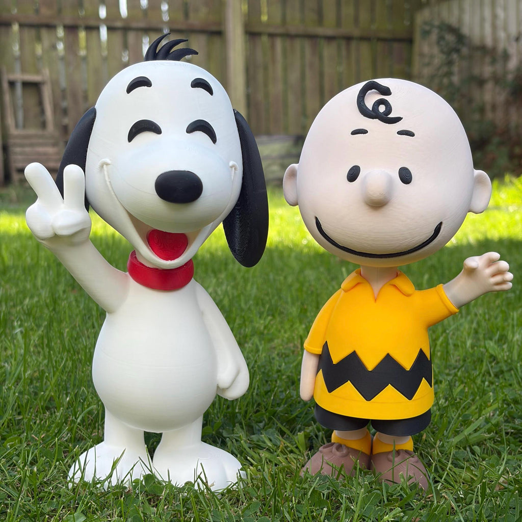 Two Peanuts character figurines, Snoopy and Charlie Brown, standing on a green grass lawn in front of a wooden fence.