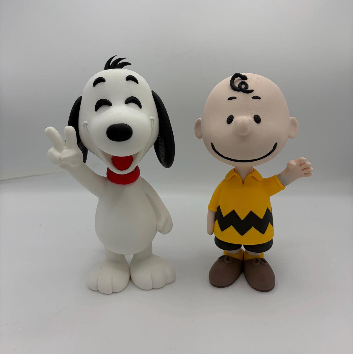 A collectible 3D printed figurine set of Snoopy and Charlie Brown from Peanuts, standing side-by-side against a neutral light grey background.