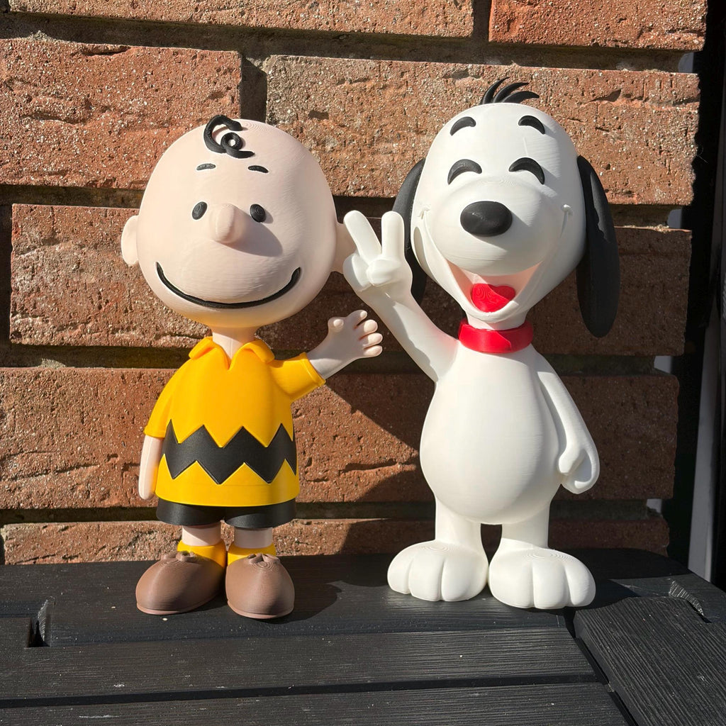 A collectible 3D printed statue of Charlie Brown and Snoopy from Peanuts, posed together on a black wooden surface in front of a red-brown brick wall.