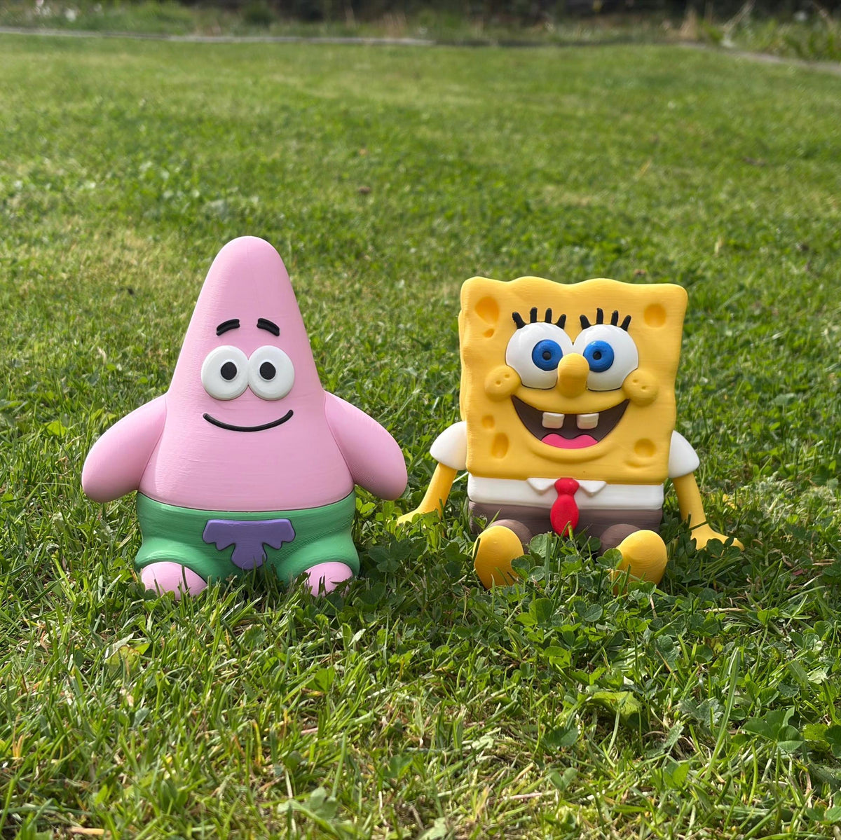 Two SpongeBob-themed toy figures, one pink star-shaped character in green shorts and one yellow square character in a white shirt with a red tie, sitting on a green grass lawn outdoors.