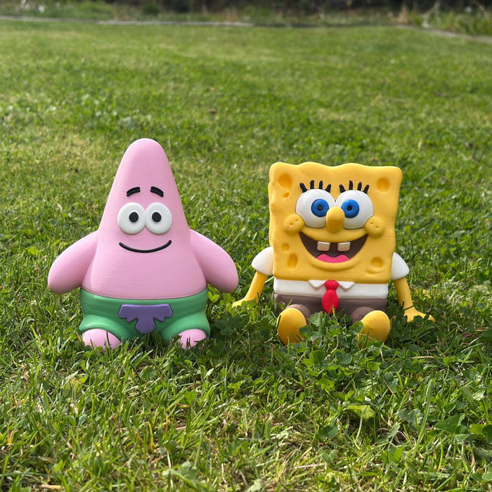 Two SpongeBob-themed toy figures, one pink star-shaped character in green shorts and one yellow square character in a white shirt with a red tie, sitting on a green grass lawn outdoors.