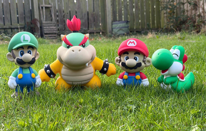 Four Super Mario character figurines, including Luigi, Bowser, Mario, and Yoshi, arranged on a green grass lawn with a wooden fence in the background.