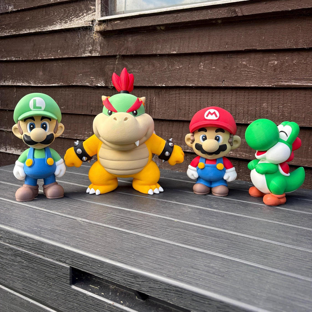 Four Super Mario character figurines lined up on a grey wooden deck in front of a brown wooden house wall. From left to right: Luigi, Bowser, Mario, and Yoshi.