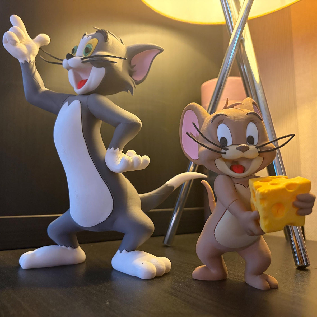 Tom and Jerry collectible figurines posed on a dark tabletop in a warmly lit indoor setting. Tom is gray and white, gesturing with one hand, while Jerry is tan holding a yellow cheese block.