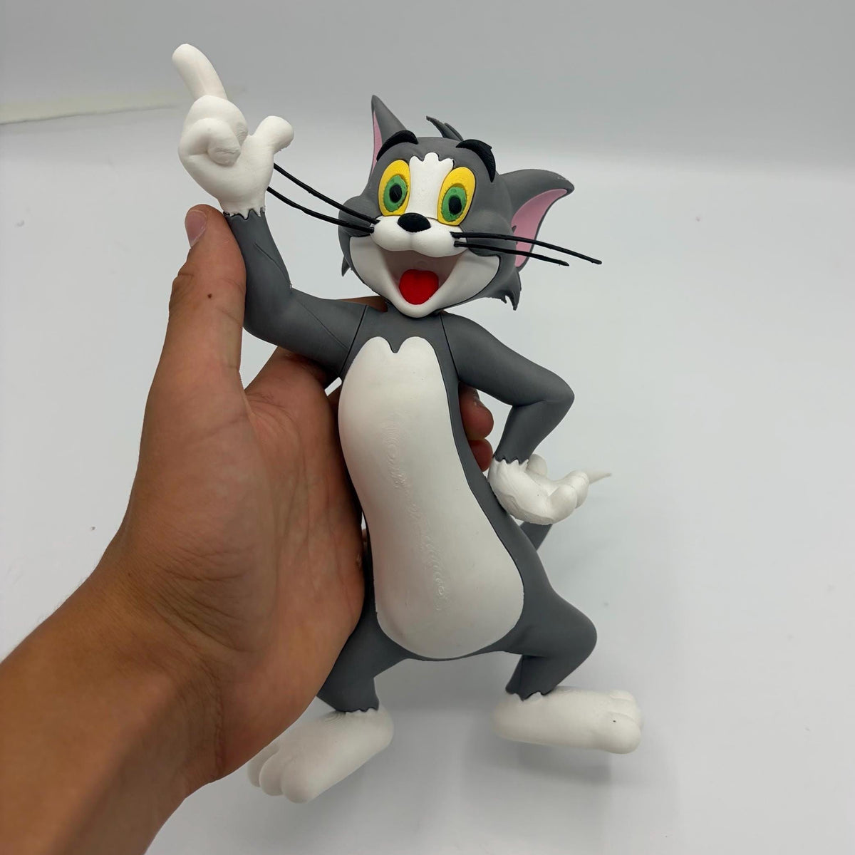 A hand holding a 3D-printed Tom and Jerry figure of Tom, the grey cat, pointing upwards. The figure has a grey and white color scheme with yellow eyes and a red mouth, set against a plain light grey background.