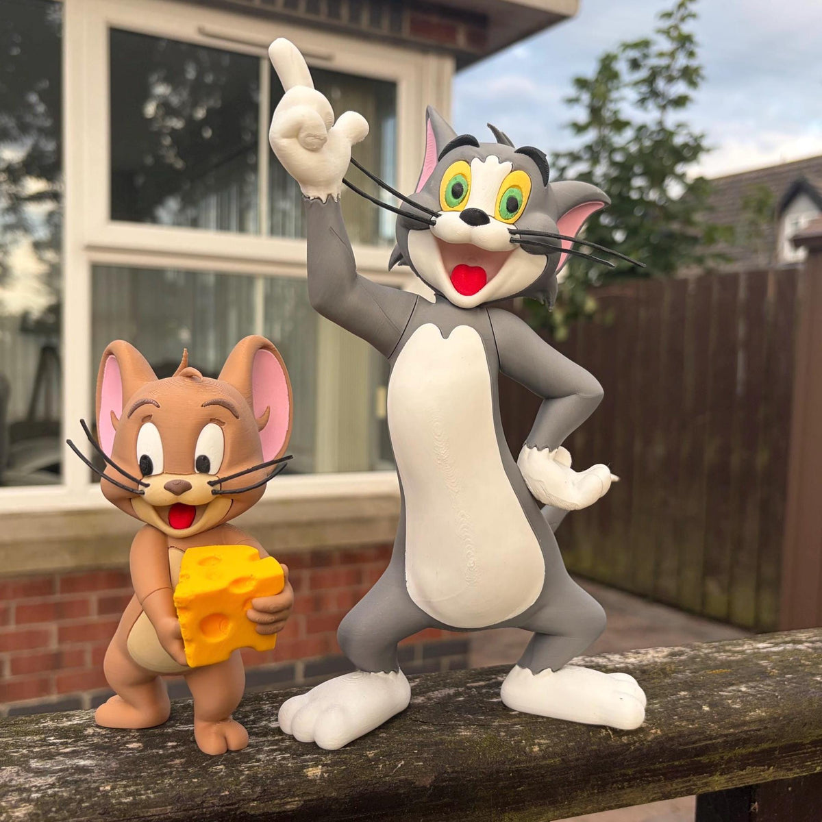 A Tom and Jerry figurine set featuring the classic grey cat Tom pointing upward and the brown mouse Jerry holding a yellow block of cheese, displayed outdoors on a wooden surface in front of a house.
