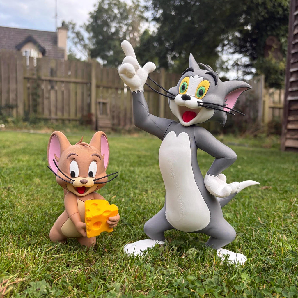 A large, detailed collectible figurine of Tom and Jerry the cartoon characters standing on a green lawn in a backyard setting. Tom is grey and white, pointing upwards, while Jerry is tan and holding a yellow cheese wedge.