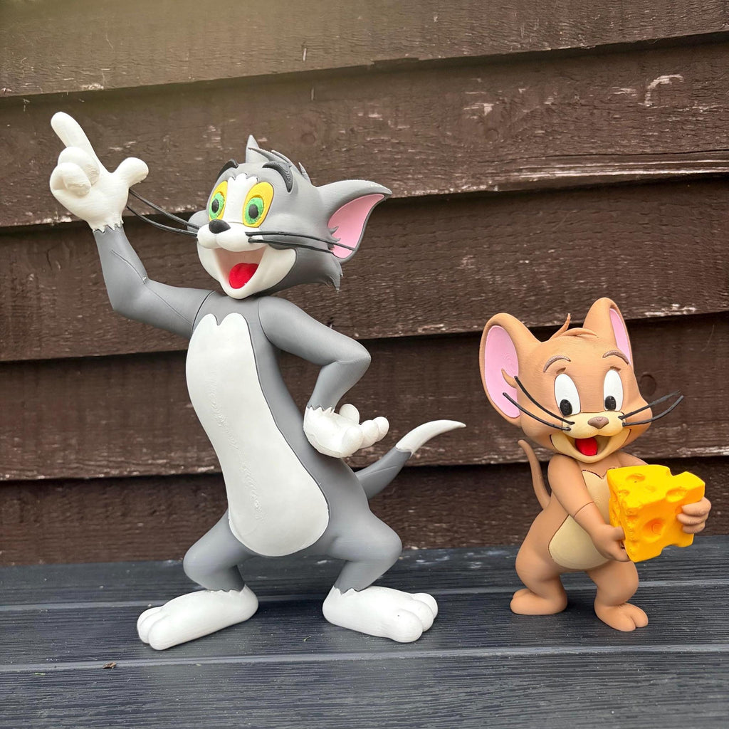 A collectible figurine set of Tom and Jerry cartoon characters standing on a dark wooden surface against a brown wooden plank background. Tom is grey and white with green eyes and is pointing upward, while Jerry is tan with pink ears and holds a yellow cheese block.