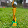 A women's FIFA World Cup trophy sculpture made of metallic gold and green plastic, standing on a green grass field with a soccer ball on its head. The base features an embossed gold FIFA logo.