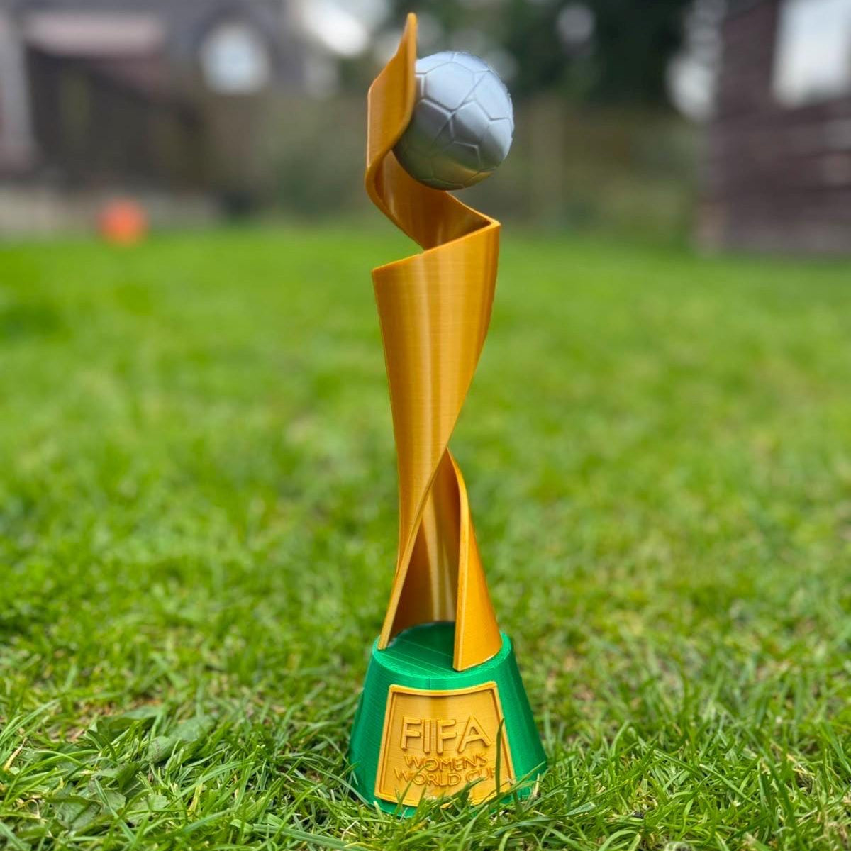 A women's FIFA World Cup trophy sculpture made of metallic gold and green plastic, standing on a green grass field with a soccer ball on its head. The base features an embossed gold FIFA logo.