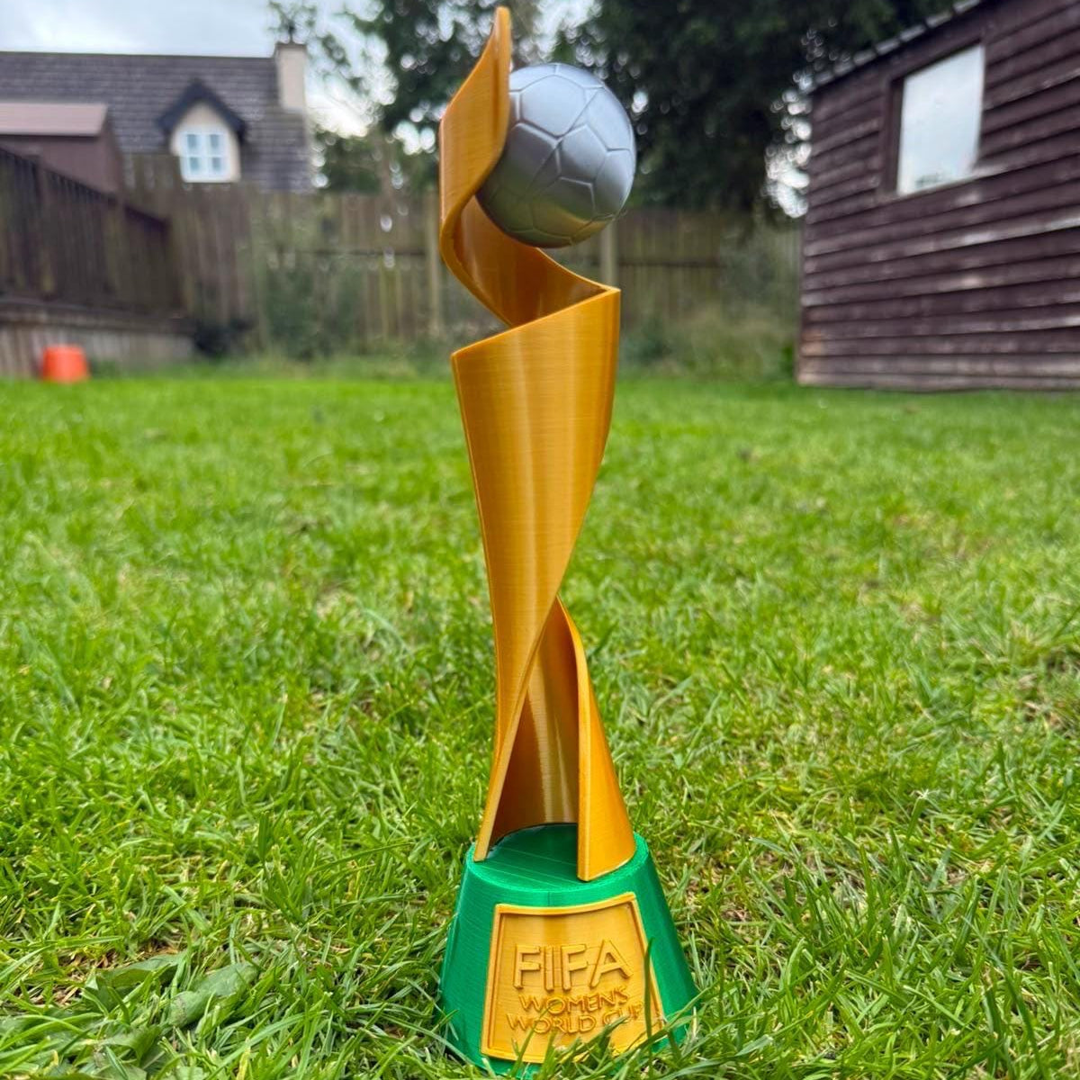 A women's FIFA World Cup trophy figurine standing on a green lawn. The figurine features a gold spiral design with a soccer ball on top and a green base engraved with 'FIFA WORLD CUP'.
