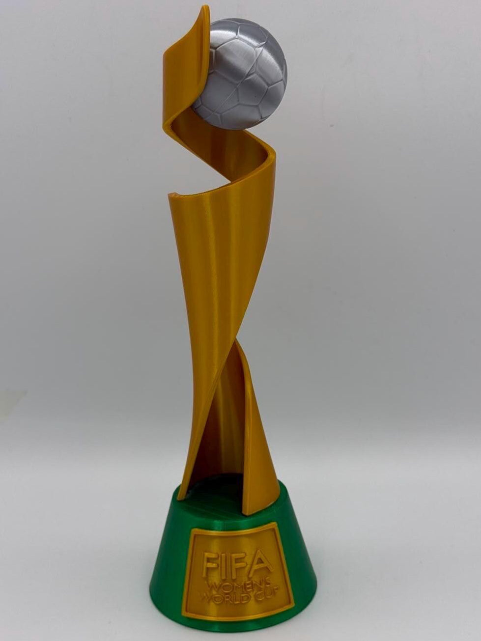 A FIFA Women's World Cup trophy replica featuring a gold spiral structure holding a silver soccer ball, mounted on a green base with a gold FIFA World Cup 1990 plaque.