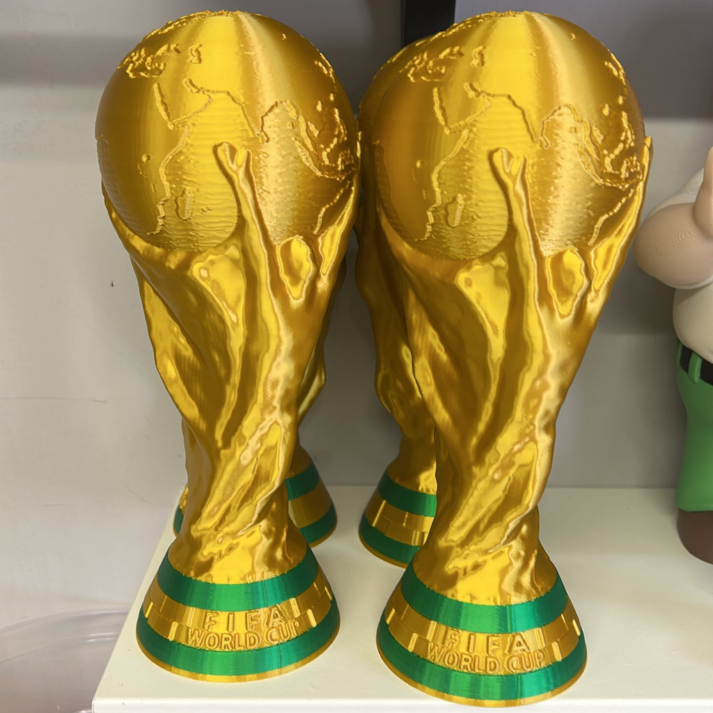 Two golden FIFA World Cup trophy replicas displayed on a white shelf, with green accents near the base.