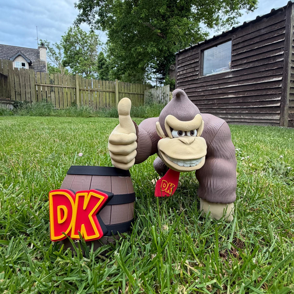 A Funko Pop! vinyl figure of Donkey Kong standing on grass in a backyard, holding a gold coin and giving a thumbs-up next to a small barrel with a red and yellow 'PK' sign.