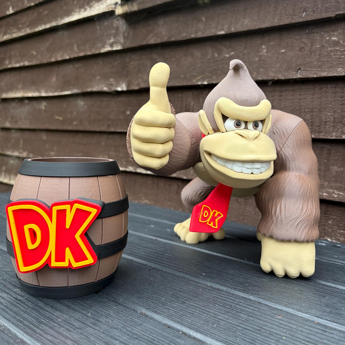 A Donkey Kong figurine standing on a dark wooden surface in front of a brown wood-paneled wall, giving a thumbs-up while wearing a red DK tie. A small brown barrel with large yellow and red DK lettering sits beside it.