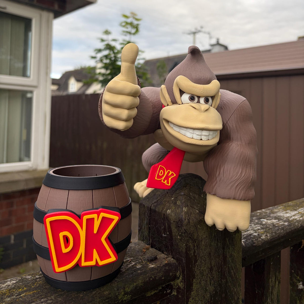 A Donkey Kong figurine giving a thumbs-up next to a small barrel-shaped mug with a large DK logo, set outdoors on a wooden fence with a residential background.
