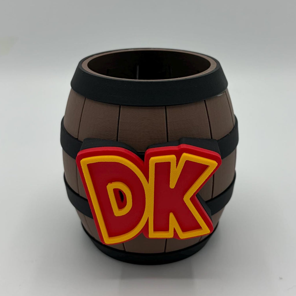 A miniature brown wooden barrel-shaped cup or mug with black bands and a red and yellow 'DK' logo on the front, set against a plain light gray background.