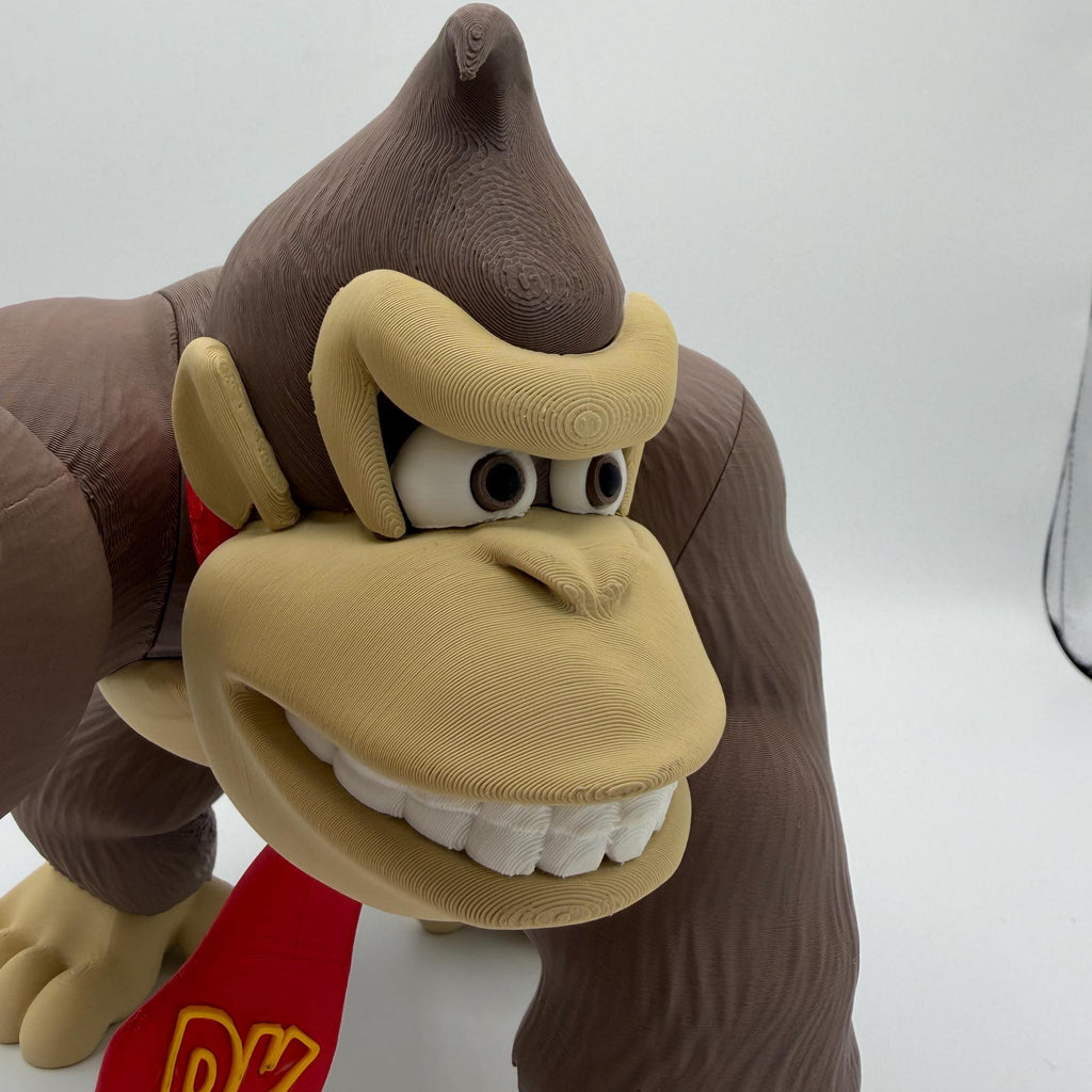 A close-up of a Donkey Kong toy figure against a plain white background, showing its brown fur, tan face, and red vest with yellow lettering.