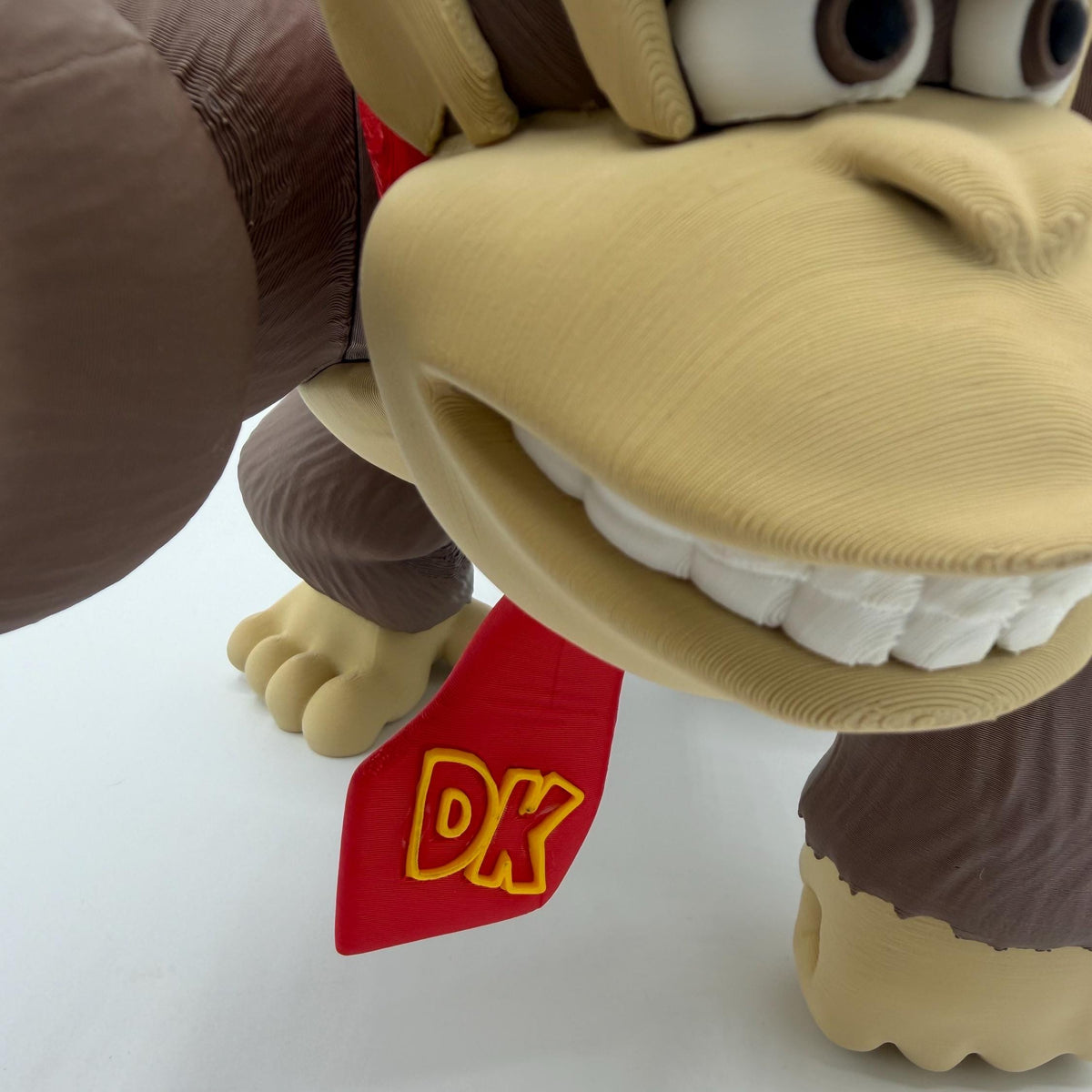 Close-up of a Donkey Kong figurine or collectible character on a white surface, showing the tan face, brown arms, white teeth, and a red neck tag with yellow 'DK' lettering.