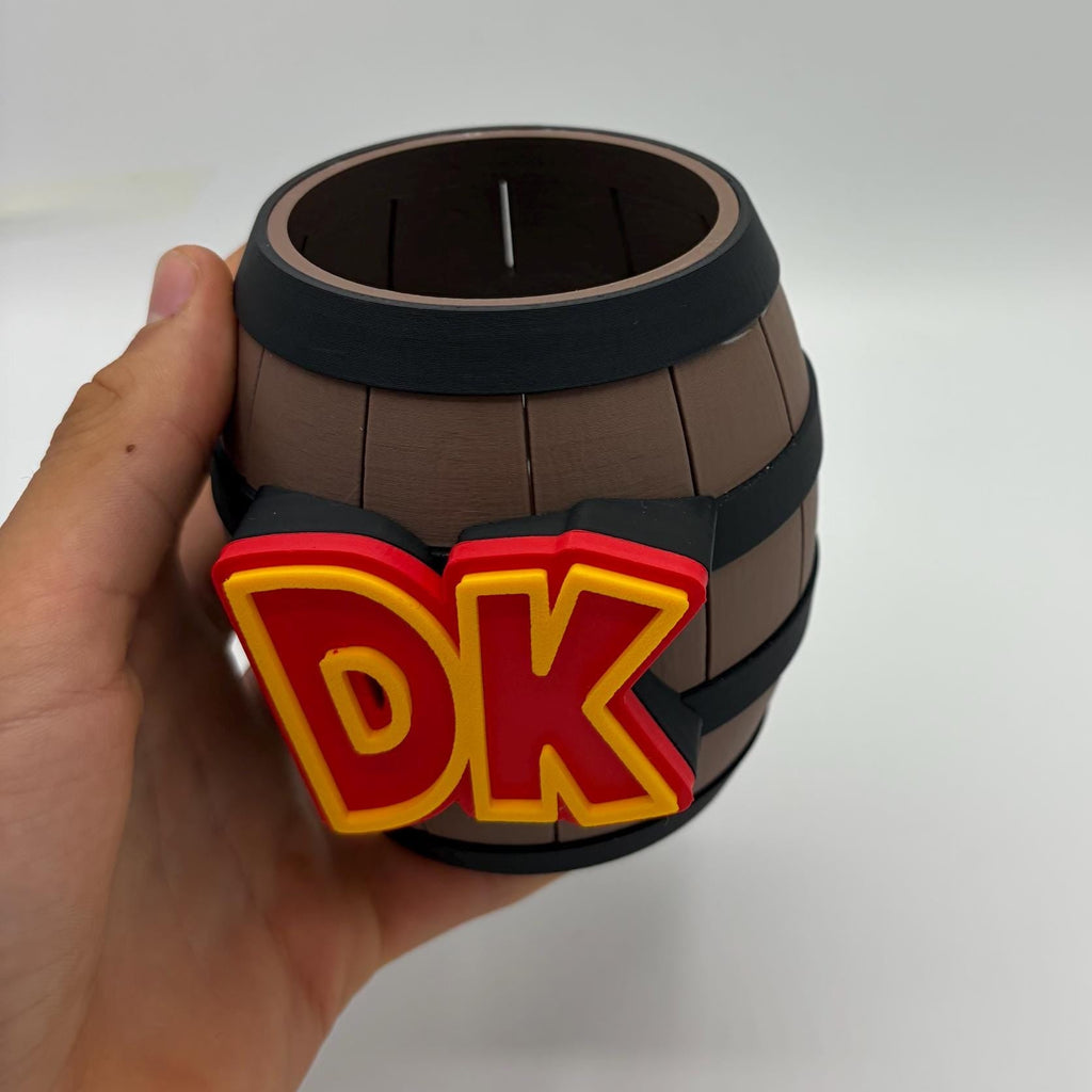 A hand holding a small barrel-shaped cup or candle holder with a brown wood-grain pattern, dark bands, and a red and yellow 'DK' emblem on the front, against a plain light gray background.
