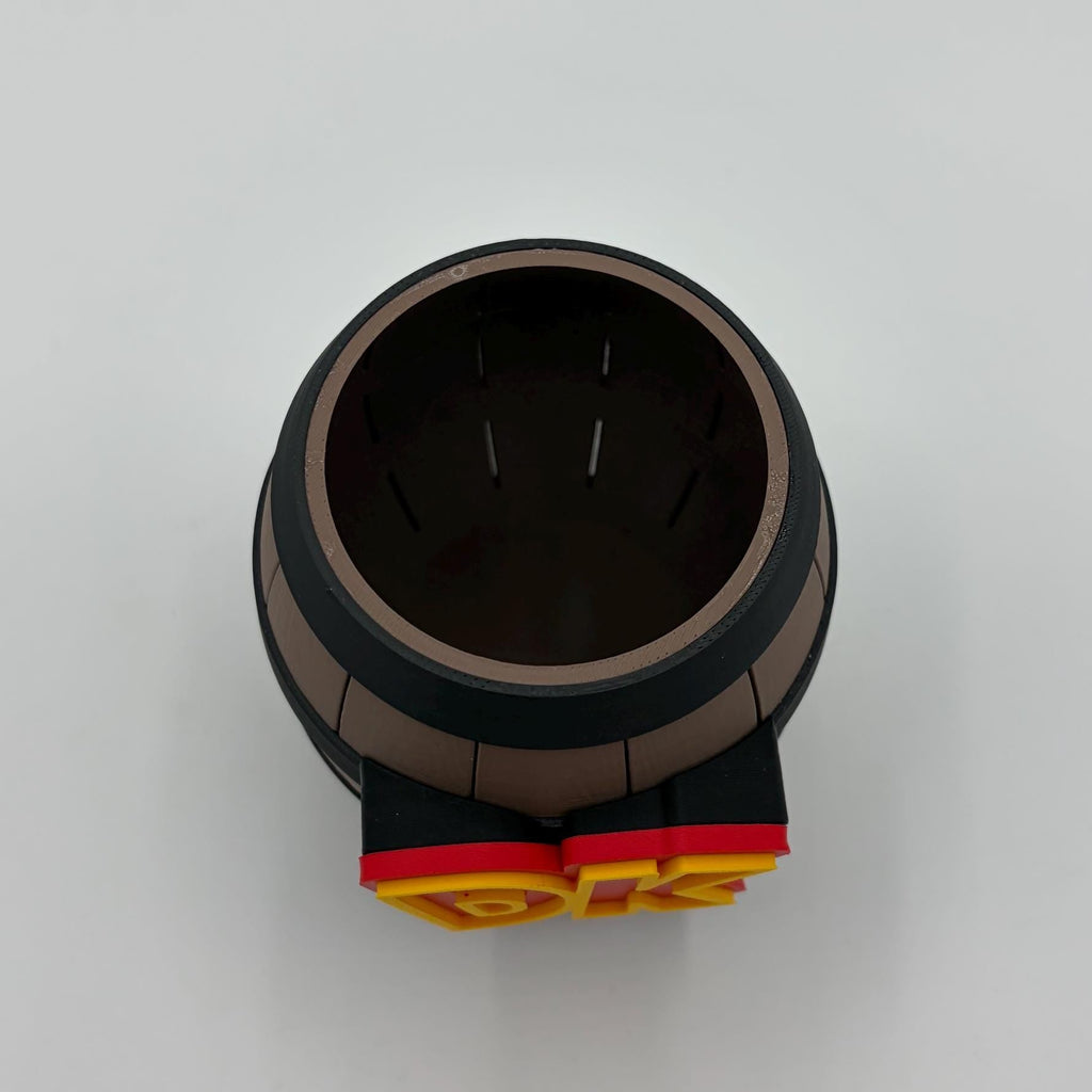 A miniature collectible model of a barrel, styled like a LEGO brick, featuring a brown and black barrel body with a red and yellow LEGO logo at the base, photographed from directly above on a light gray background.