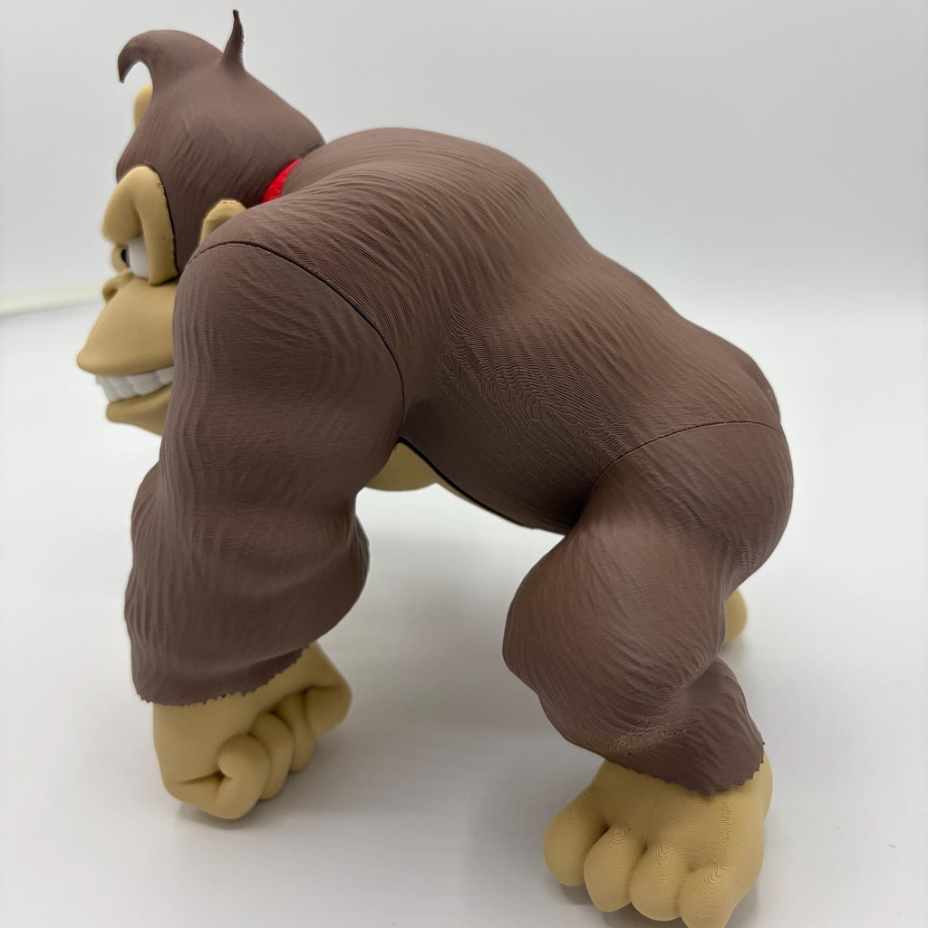 A plastic toy figure of a brown gorilla in a crouching, forward-facing pose, featuring tan skin on its face, hands, and feet, and a small red collar, set against a plain white studio background.
